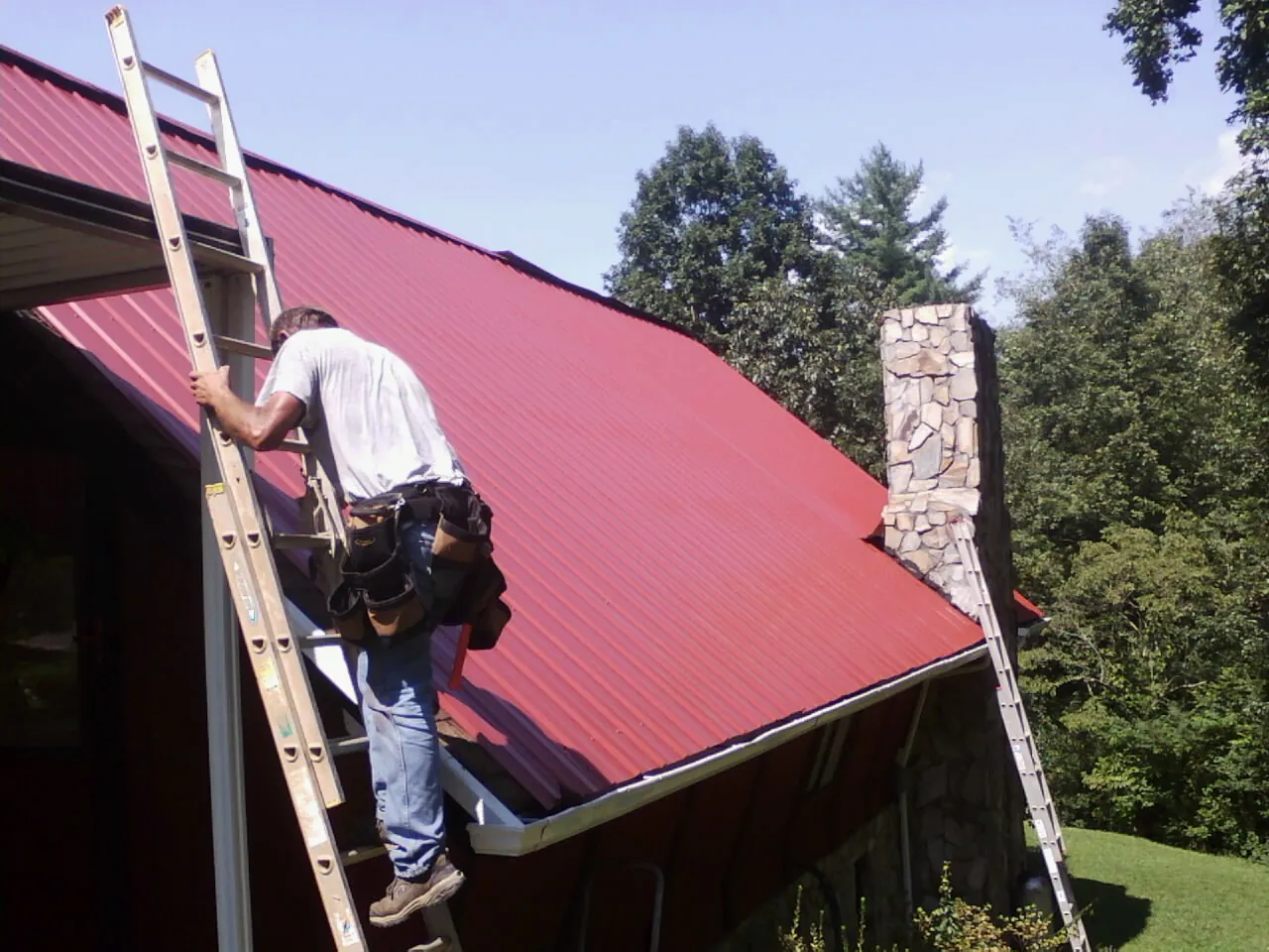 Roofer on a ladder beside a red metal roof and stone chimney — Soffit & Fascia Repair by Roofing Brothers in Grindstone