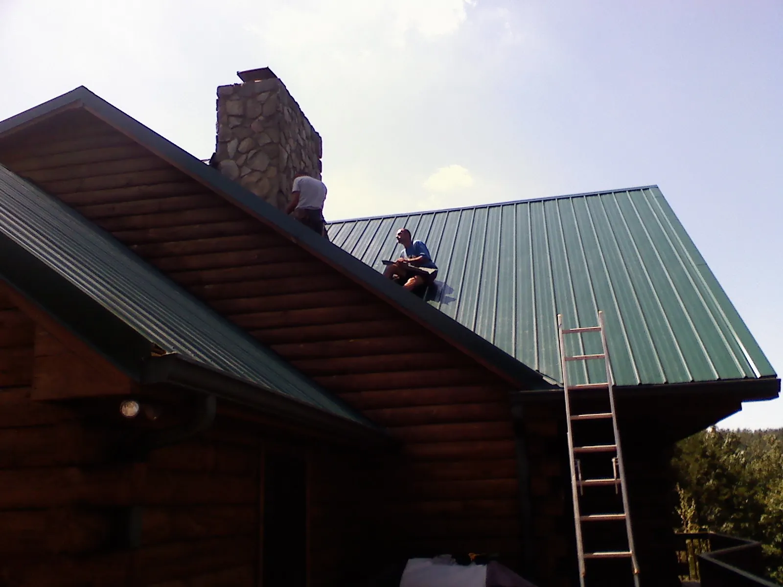 Roofers installing green metal panels beside a stone chimney on a log home in Grindstone